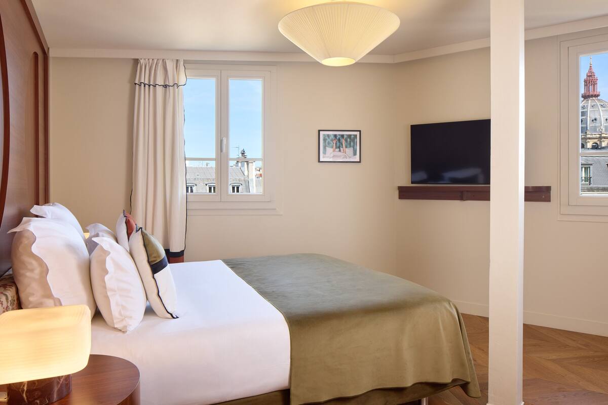 A hotel room with a bed, a television, a window with curtains, and a view of a building.