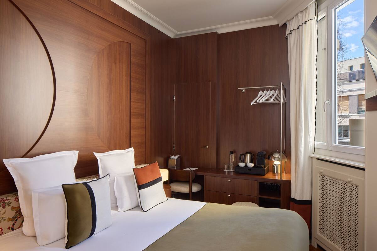 A hotel room with a bed, bedside table, desk, chair, and a window with curtains.
