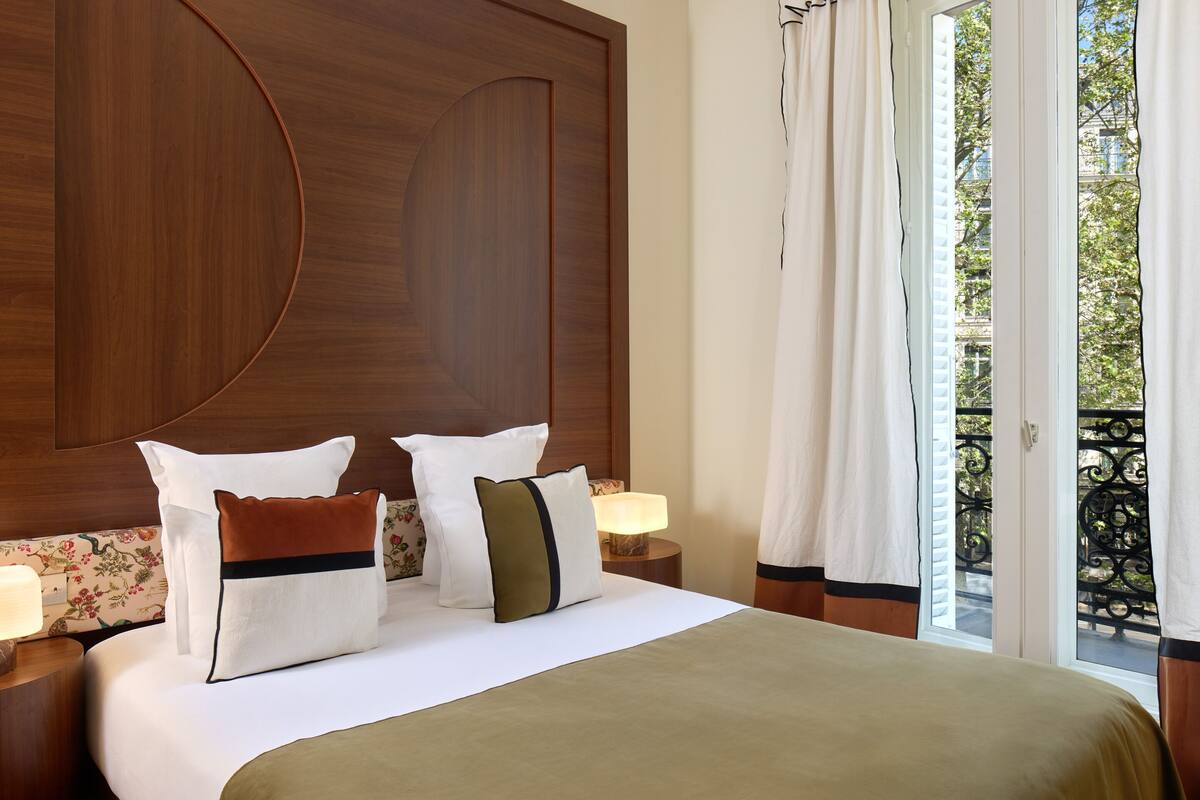 A hotel room with a large bed, a wooden headboard, a nightstand with a lamp, and a view of a balcony.
