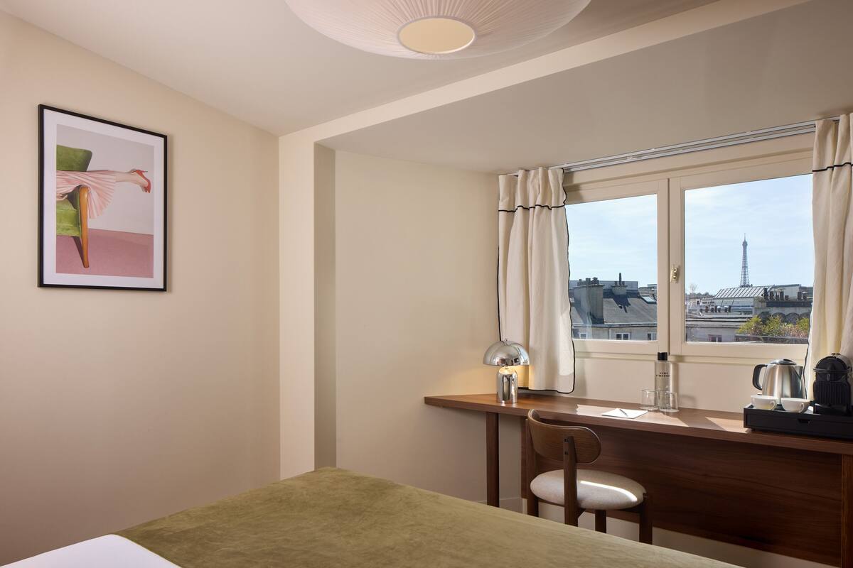A hotel room with a bed, a desk with a lamp, a chair, a window with curtains, and a view of a tower.