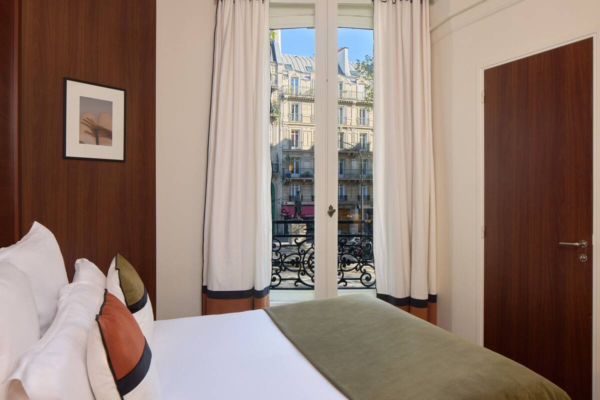 A hotel room with a bed, curtains, a framed picture, and a view of buildings through a window.