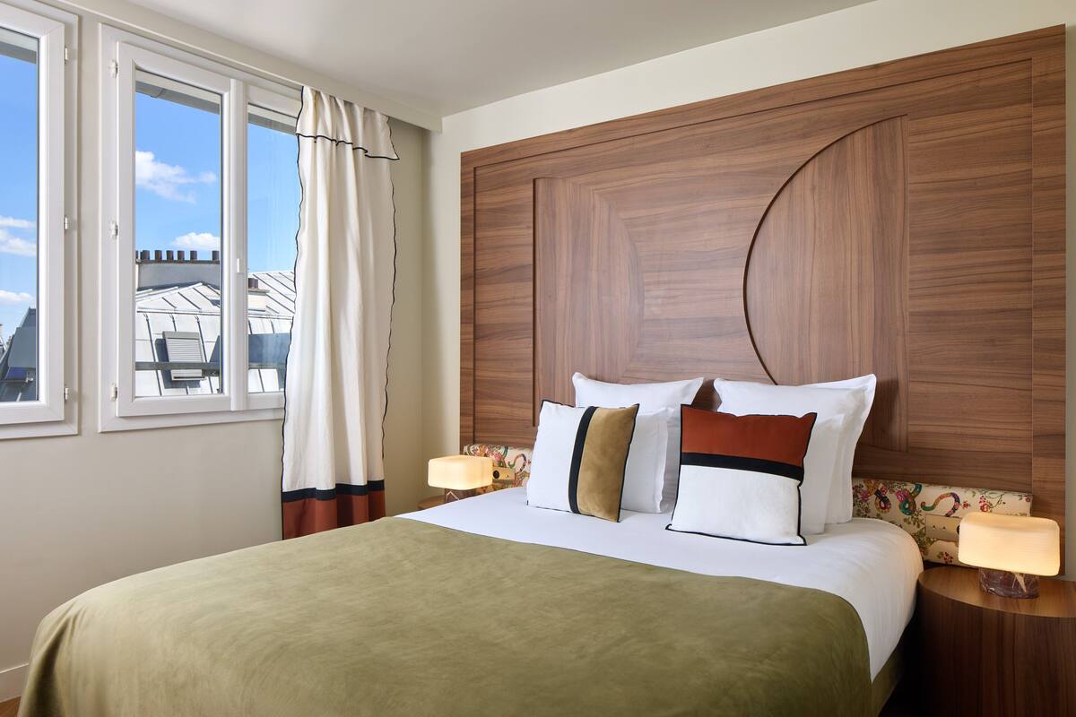 A hotel room with a bed, a nightstand, a lamp, a window with curtains, and a view of buildings outside.