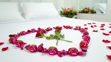 A bed with a heart-shaped arrangement of pink candies and a single rose, surrounded by scattered rose petals.
