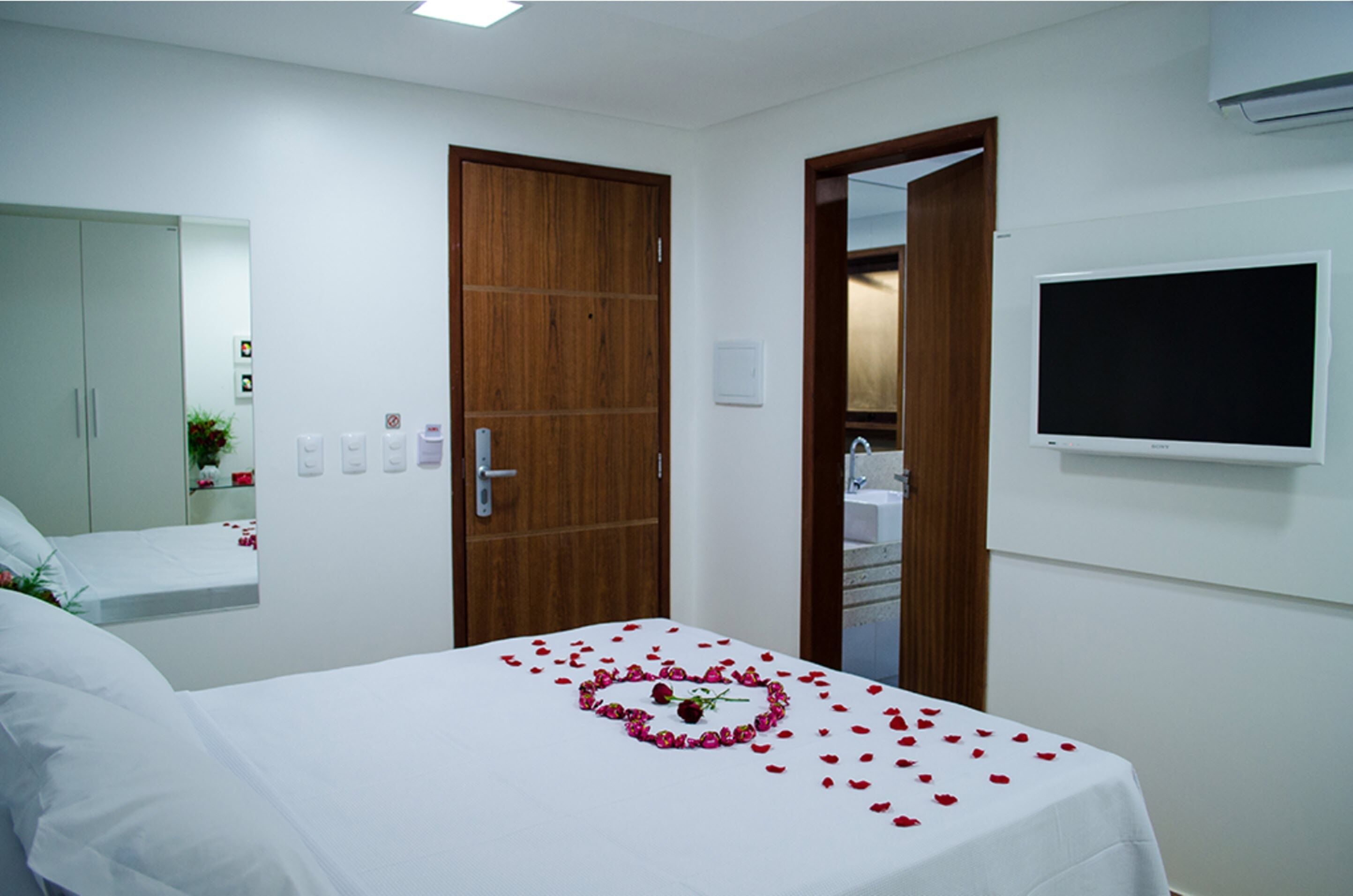 A hotel room with a bed, a wooden door, a white wall-mounted TV, and a bathroom visible through an open door.
