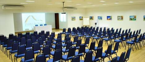 Un salón de conferencias con filas de sillas azules, un atril y una pantalla para presentaciones.