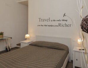Een opgemaakt bed met een gestreept dekbedovertrek, een muur met een reisquote en nachtlampjes.