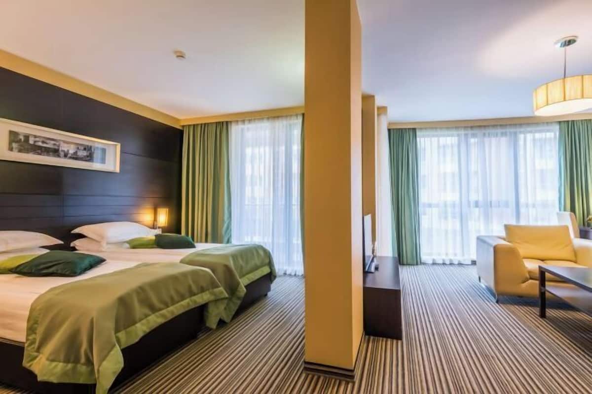 A hotel room with two beds, a sofa, a desk, and a large window with curtains.