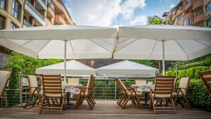Un patio al aire libre con sillas de madera y grandes sombrillas blancas, rodeado de follaje y un edificio al fondo.