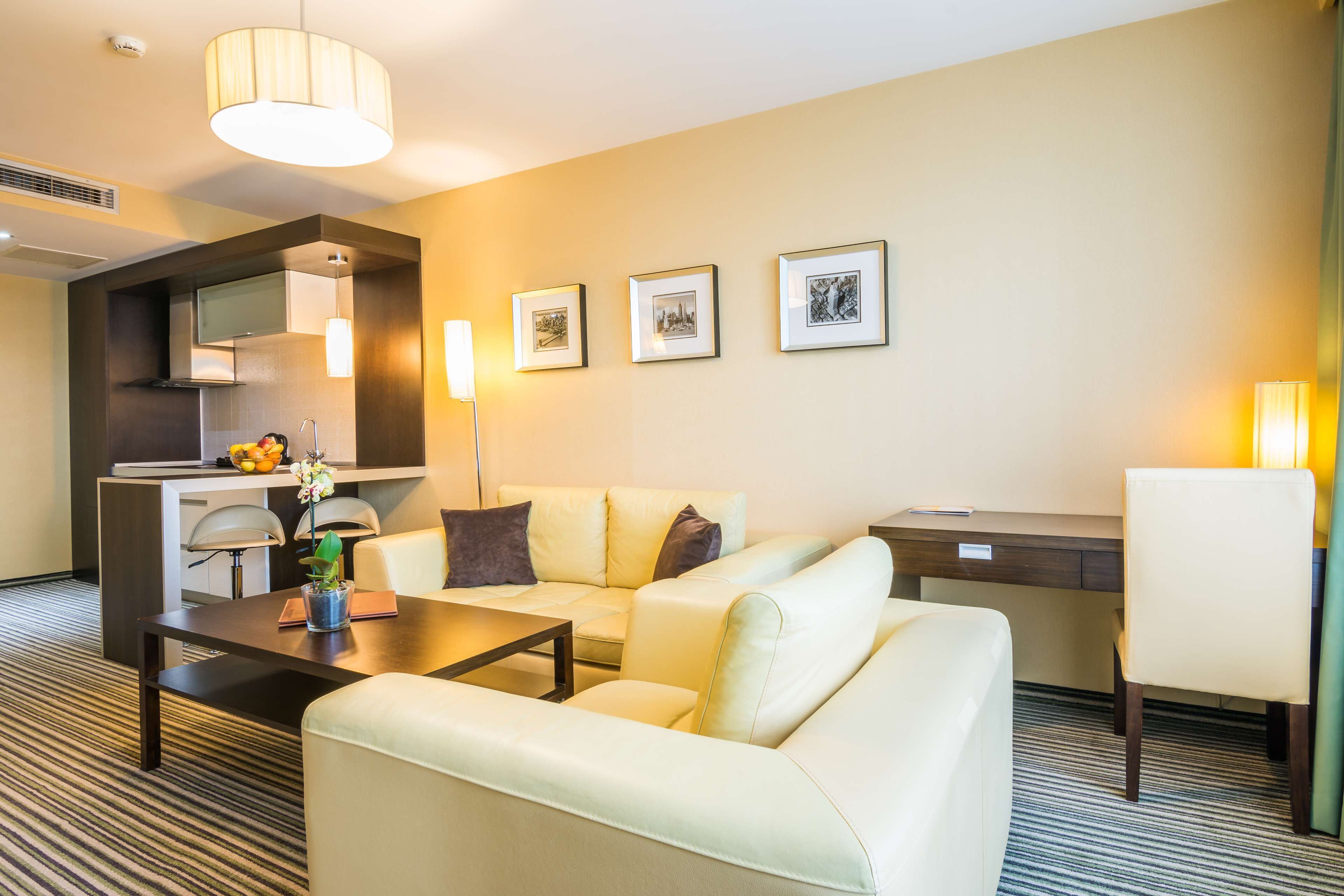 Quarto de hotel com sofá, mesa de jantar, escrivaninha, cozinha compacta e quadros nas paredes.