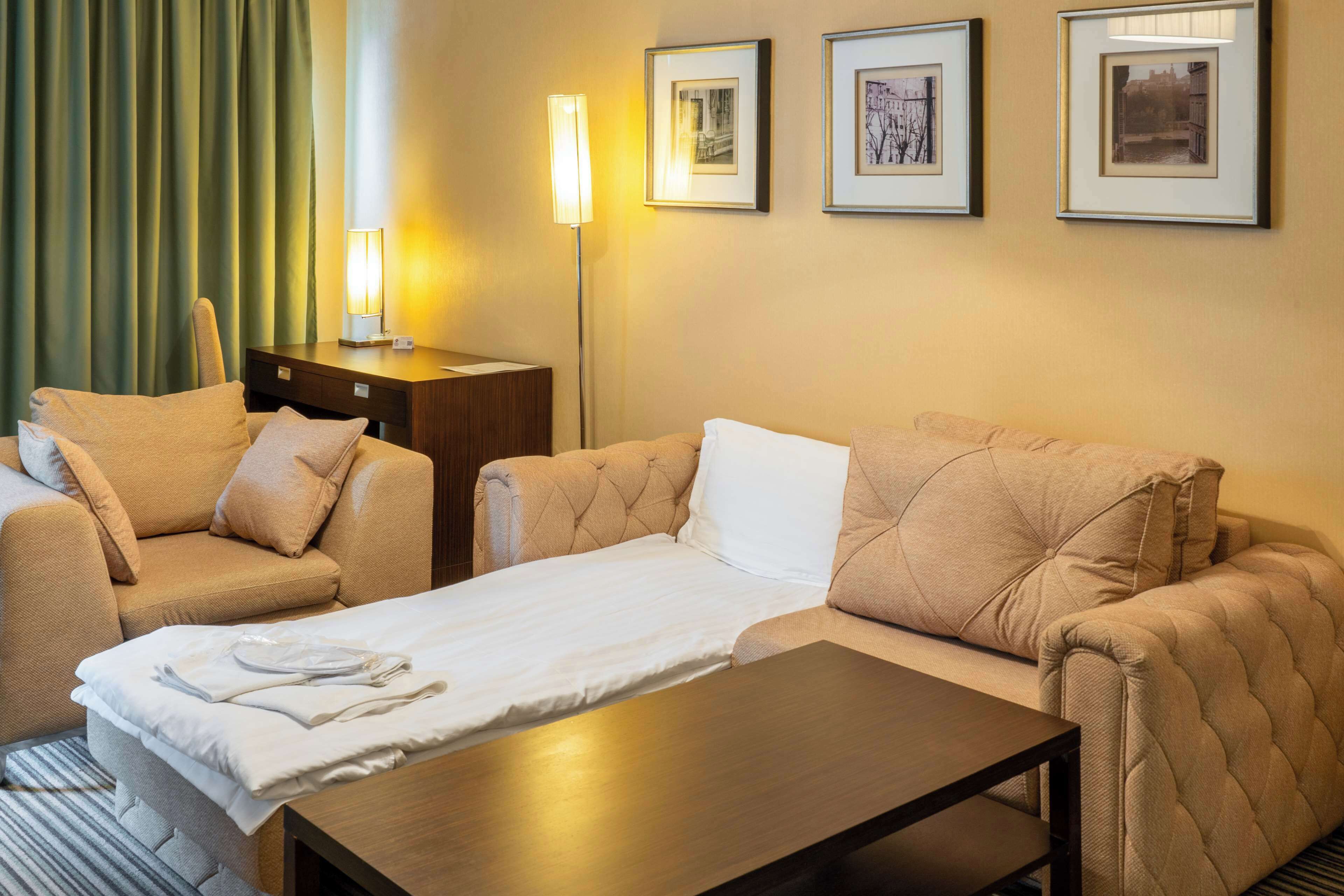 A hotel room with a bed, two armchairs, a small table, and a nightstand. There are three framed pictures on the wall.