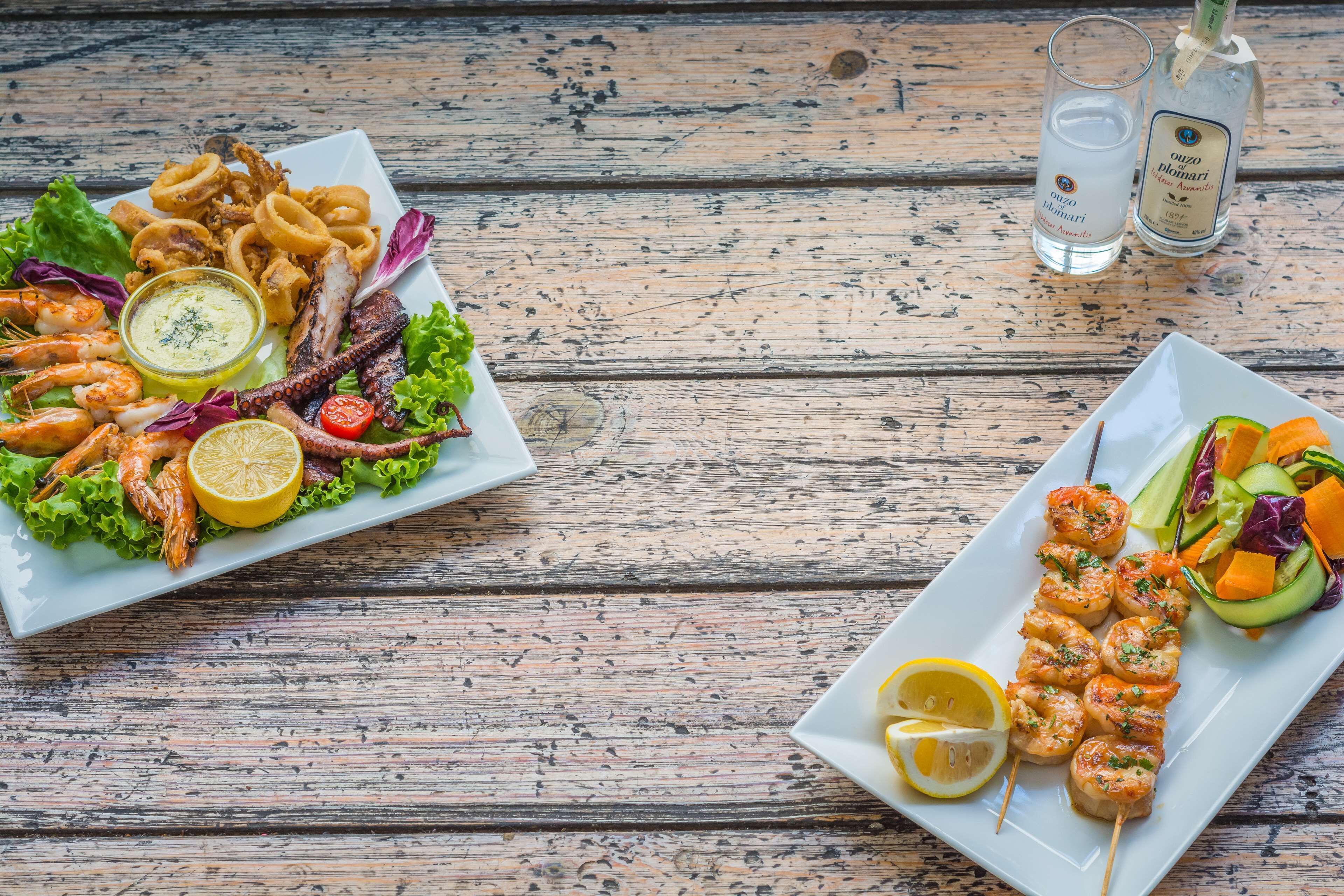 A wooden table with two plates of seafood, including shrimp, grilled octopus, and grilled squid, along with a salad and a bottle of olive oil.