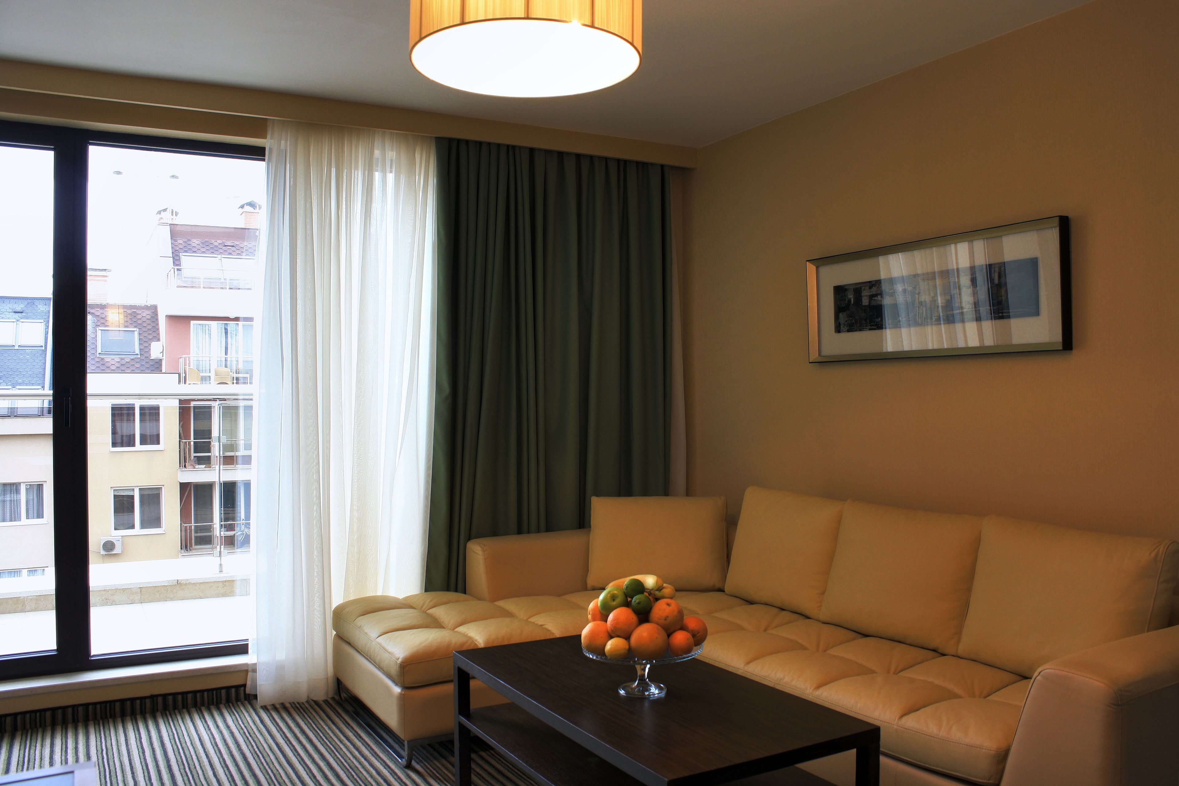 Habitación de hotel con sofá, una mesa de centro con fruta, ventana con cortinas y un cuadro en la pared.