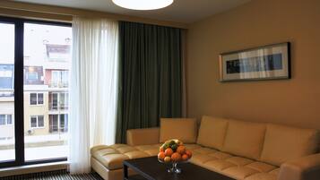Habitación de hotel con sofá, una mesa de centro con fruta, ventana con cortinas y un cuadro en la pared.