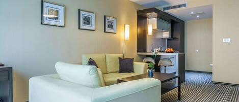 A hotel room with a sofa, a coffee table, a lamp, framed pictures on the wall, and a kitchen area with a sink and countertop.