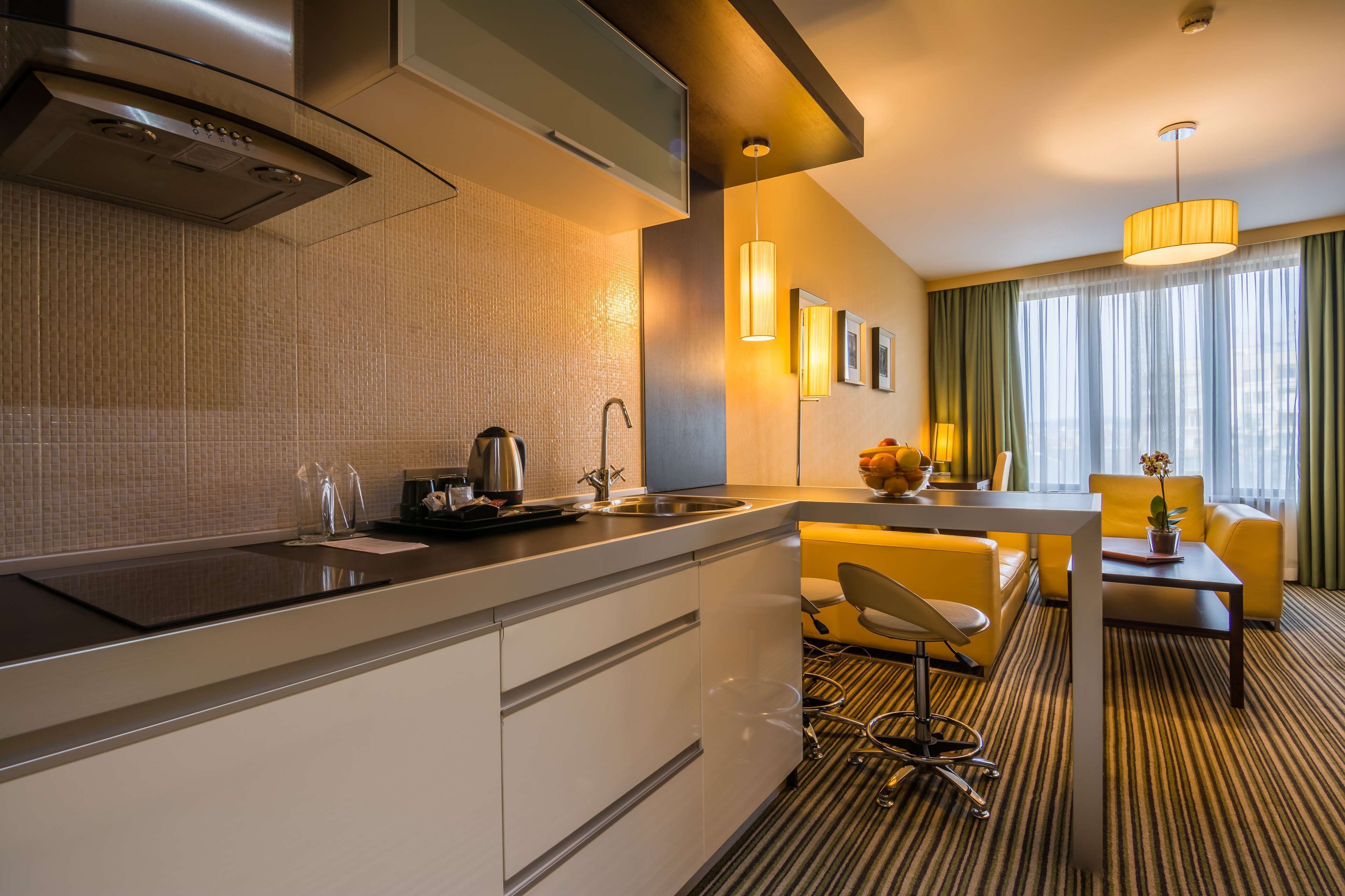 A modern hotel room with a kitchen area, a dining table with yellow chairs, and a large window with curtains.