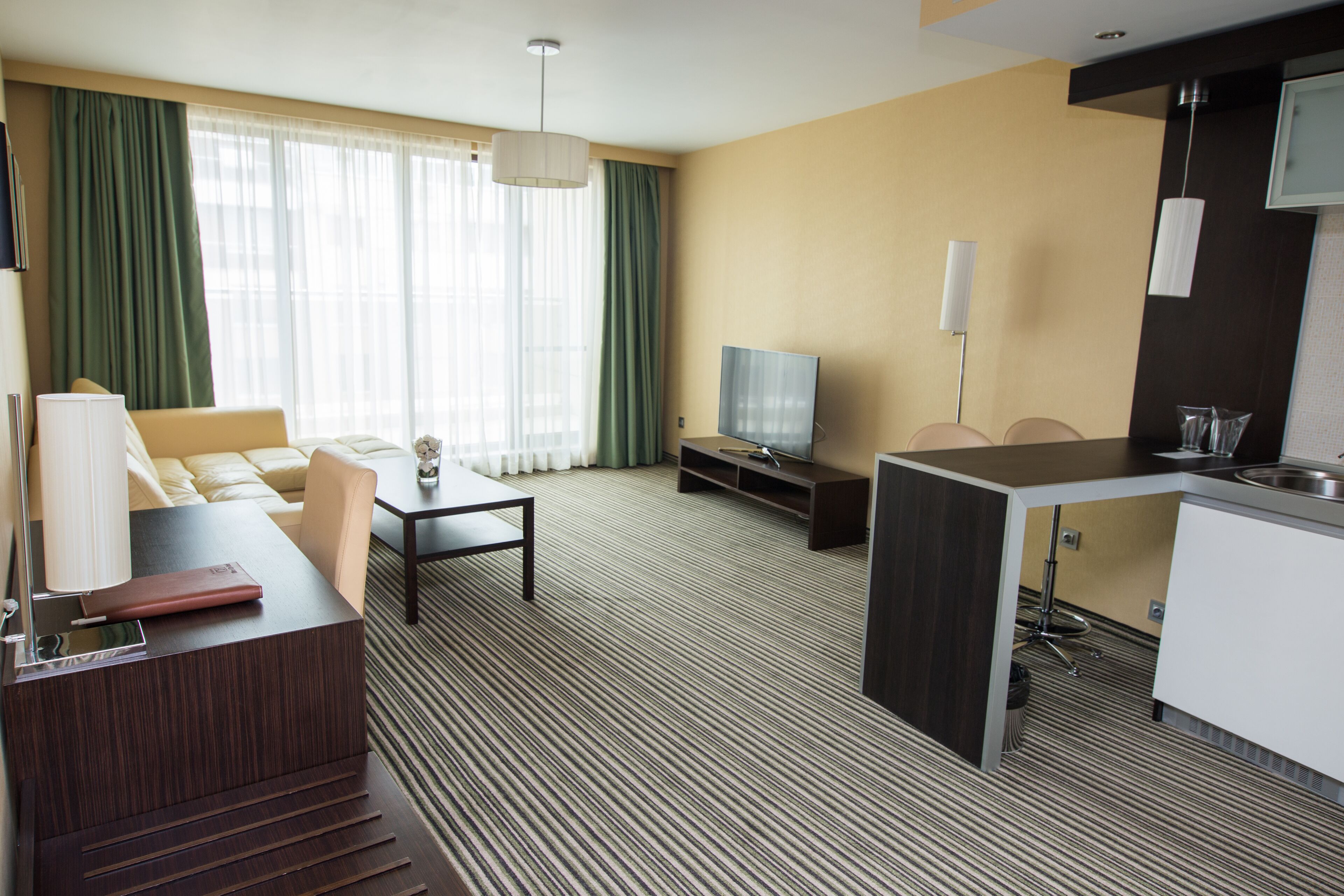 Et hotelværelse med sofa, lille sofabord, fjernsyn, tekøkken og et stort vindue med gardiner.