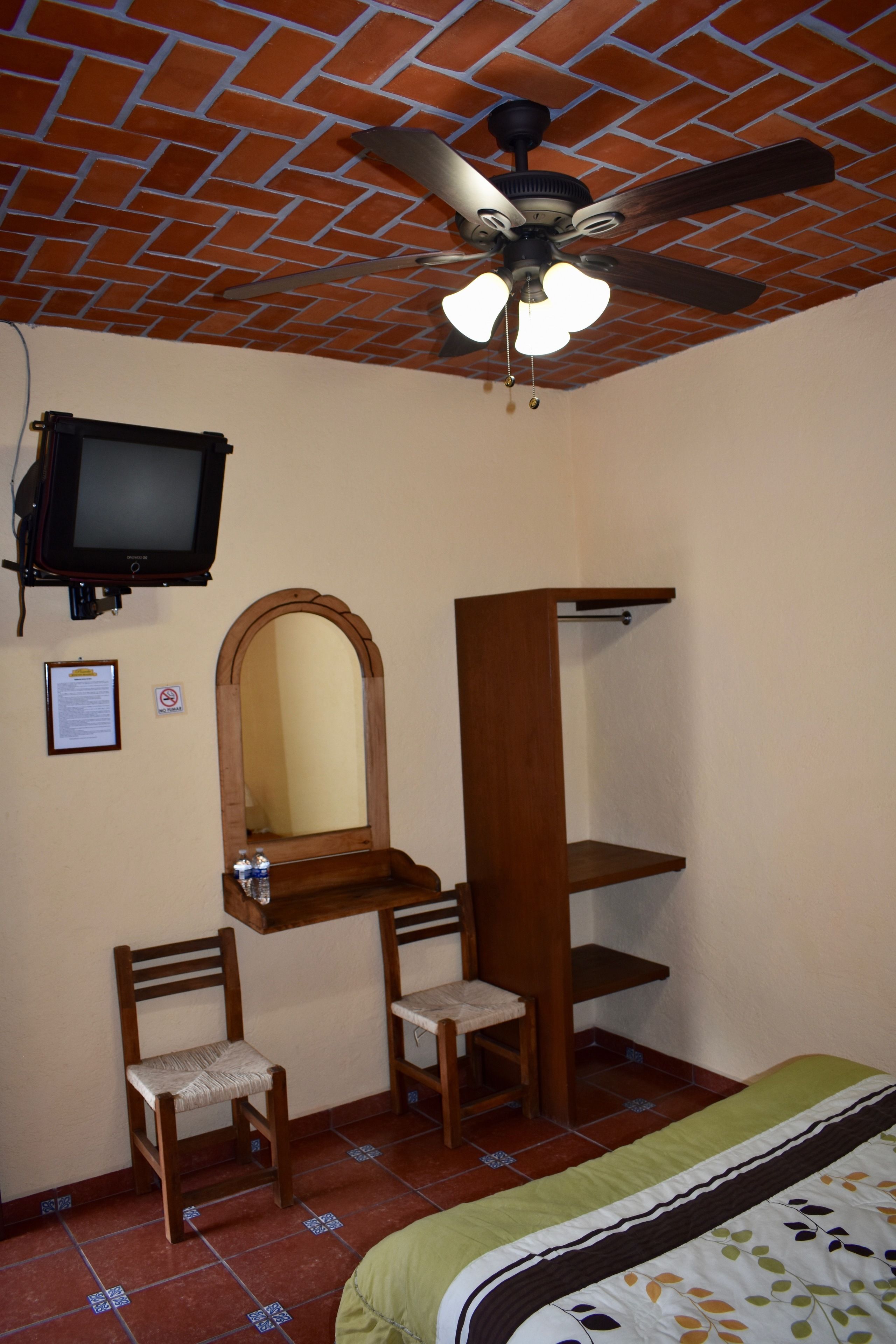 A bedroom with a brick ceiling, a ceiling fan, a television, a mirror, and a bed with a green and white bedspread.