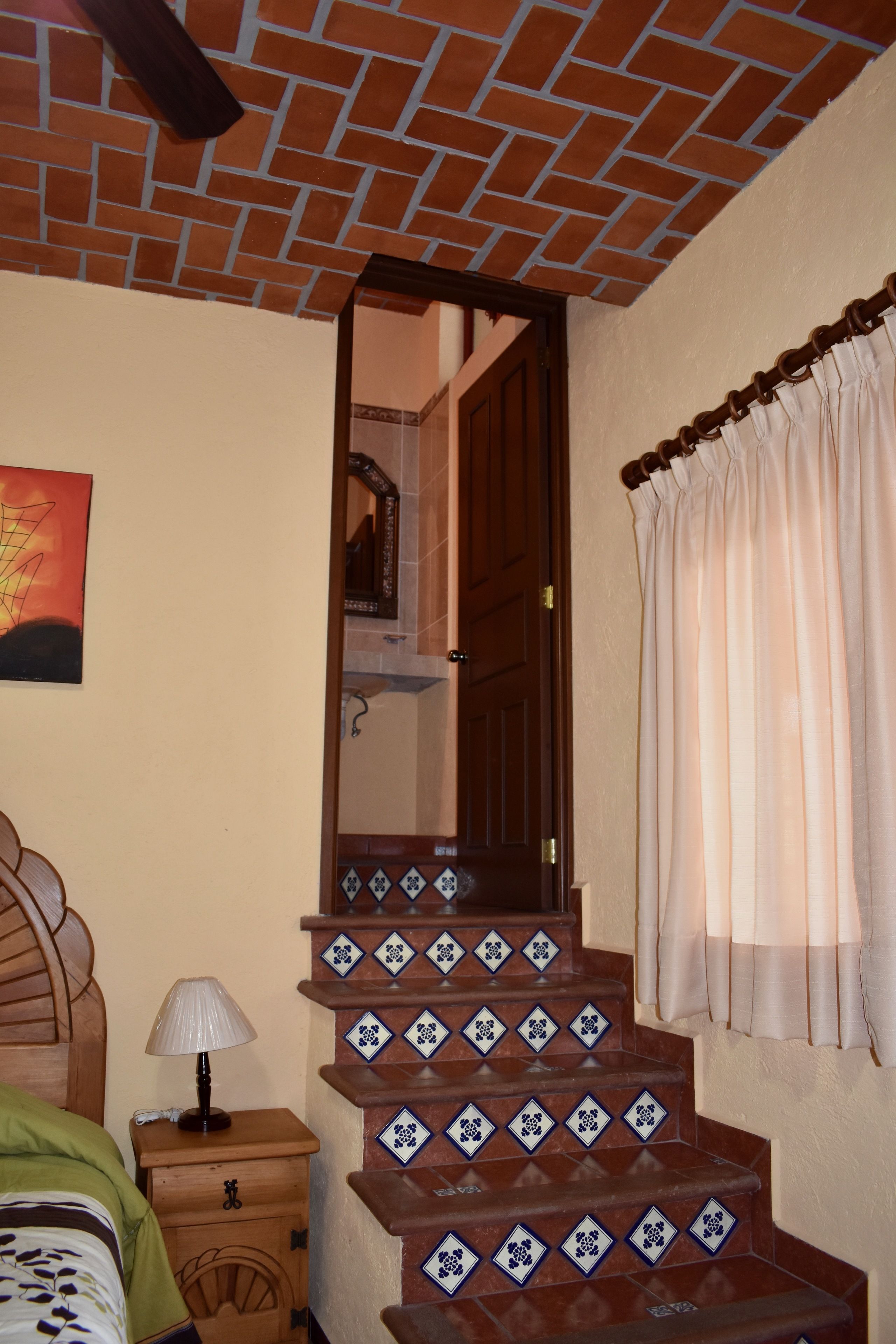 Un escalier recouvert de carreaux bleus et blancs formant un motif, une table de chevet en bois avec une lampe, et un lit avec une couverture verte.