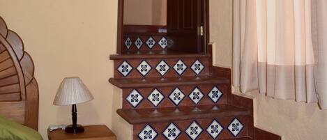 Un escalier recouvert de carreaux bleus et blancs formant un motif, une table de chevet en bois avec une lampe, et un lit avec une couverture verte.