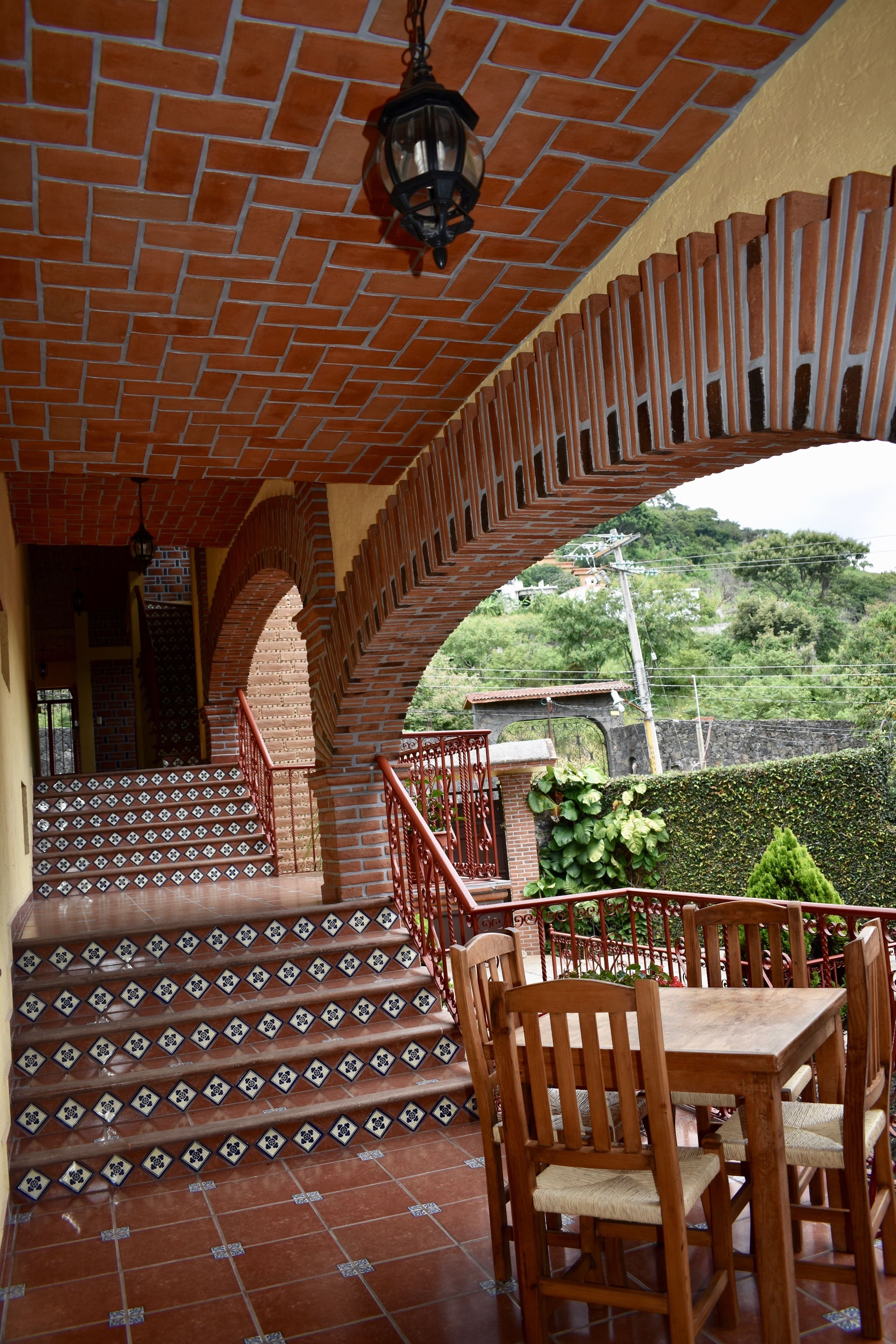 A covered outdoor area with a brick staircase leading to a higher level, wooden furniture, and a view of greenery in the background.