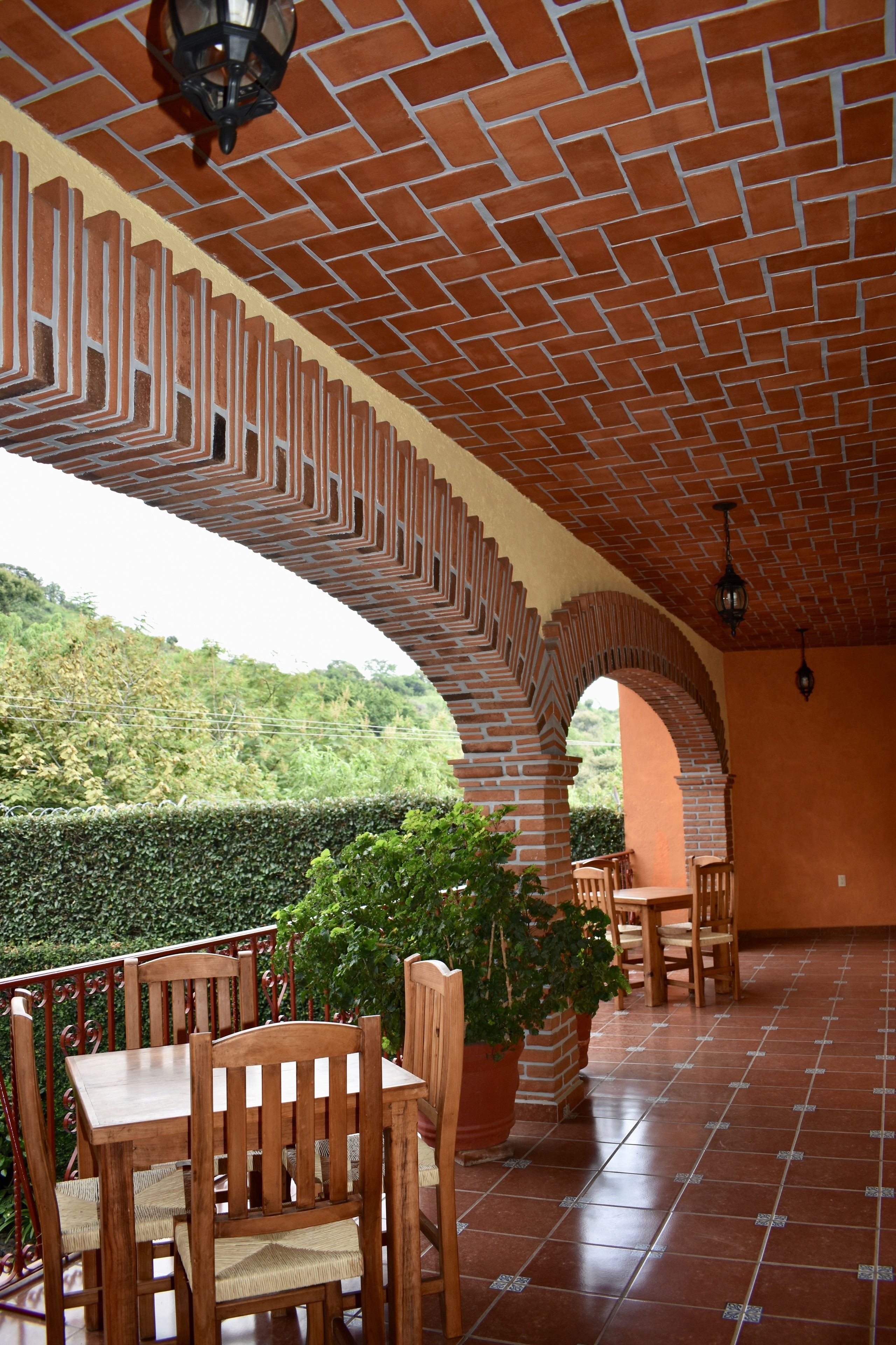 A covered patio with brick arches, wooden furniture, and potted plants.