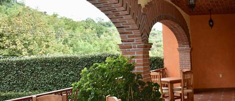 A covered patio with brick arches, wooden furniture, and potted plants.