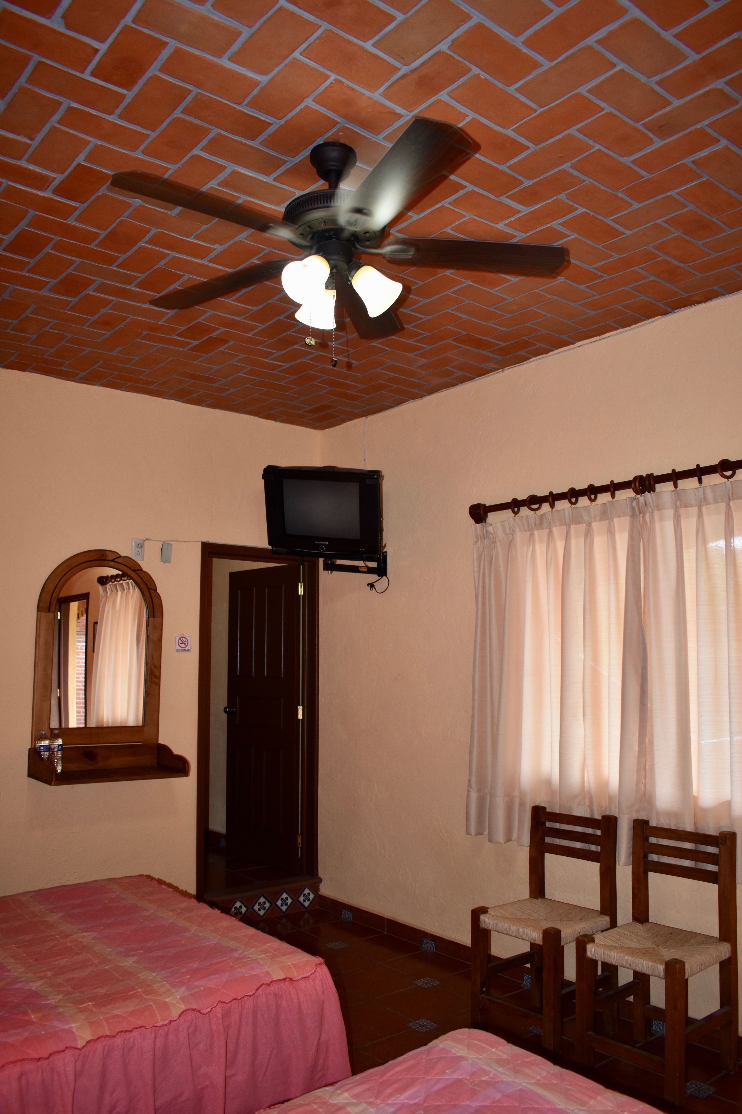 A room with two beds, a ceiling fan, a television, a mirror, and a window with curtains.