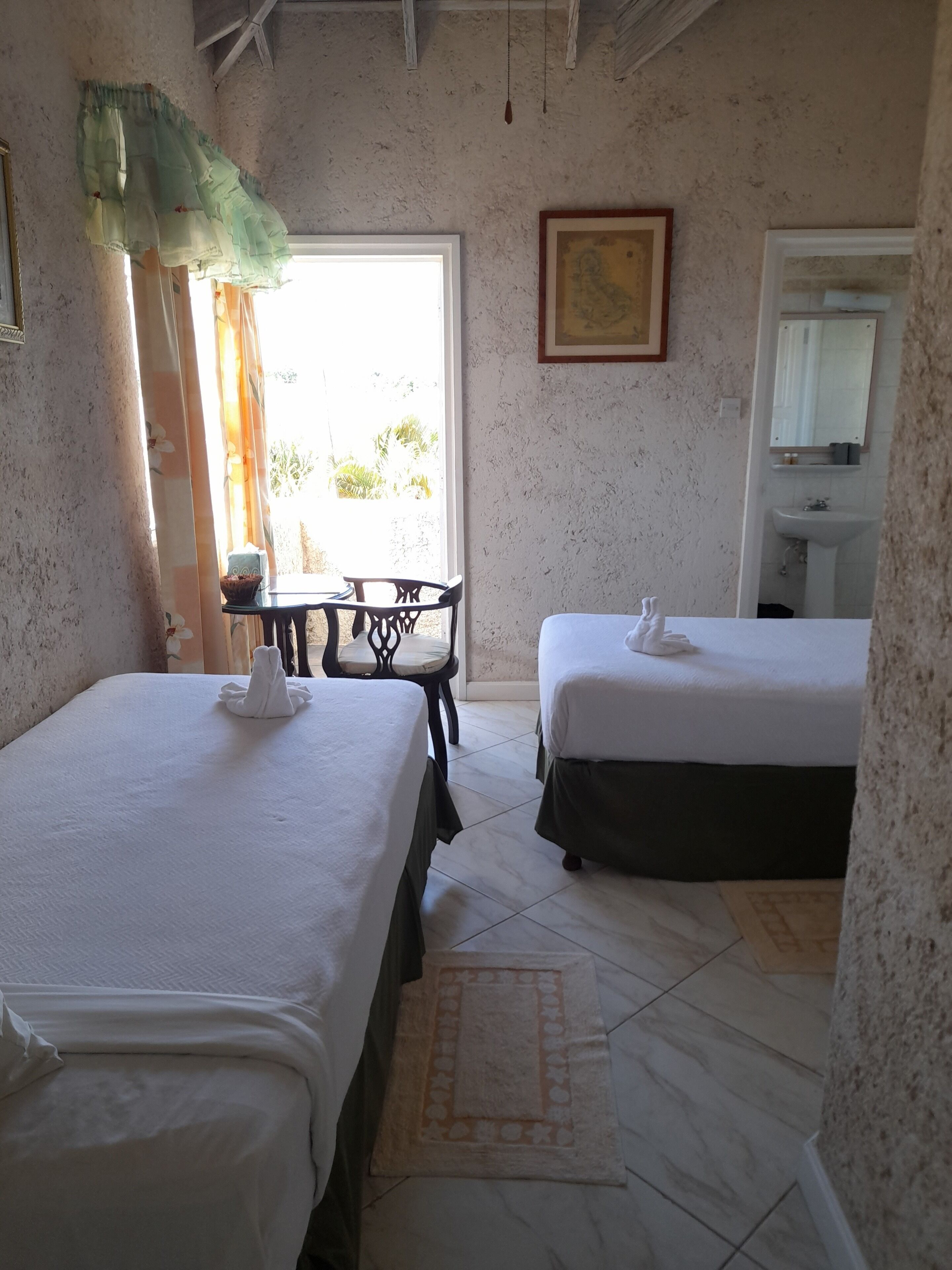 Une chambre avec deux lits, une petite table, une chaise, un tableau et une salle de bain avec un lavabo et un miroir.