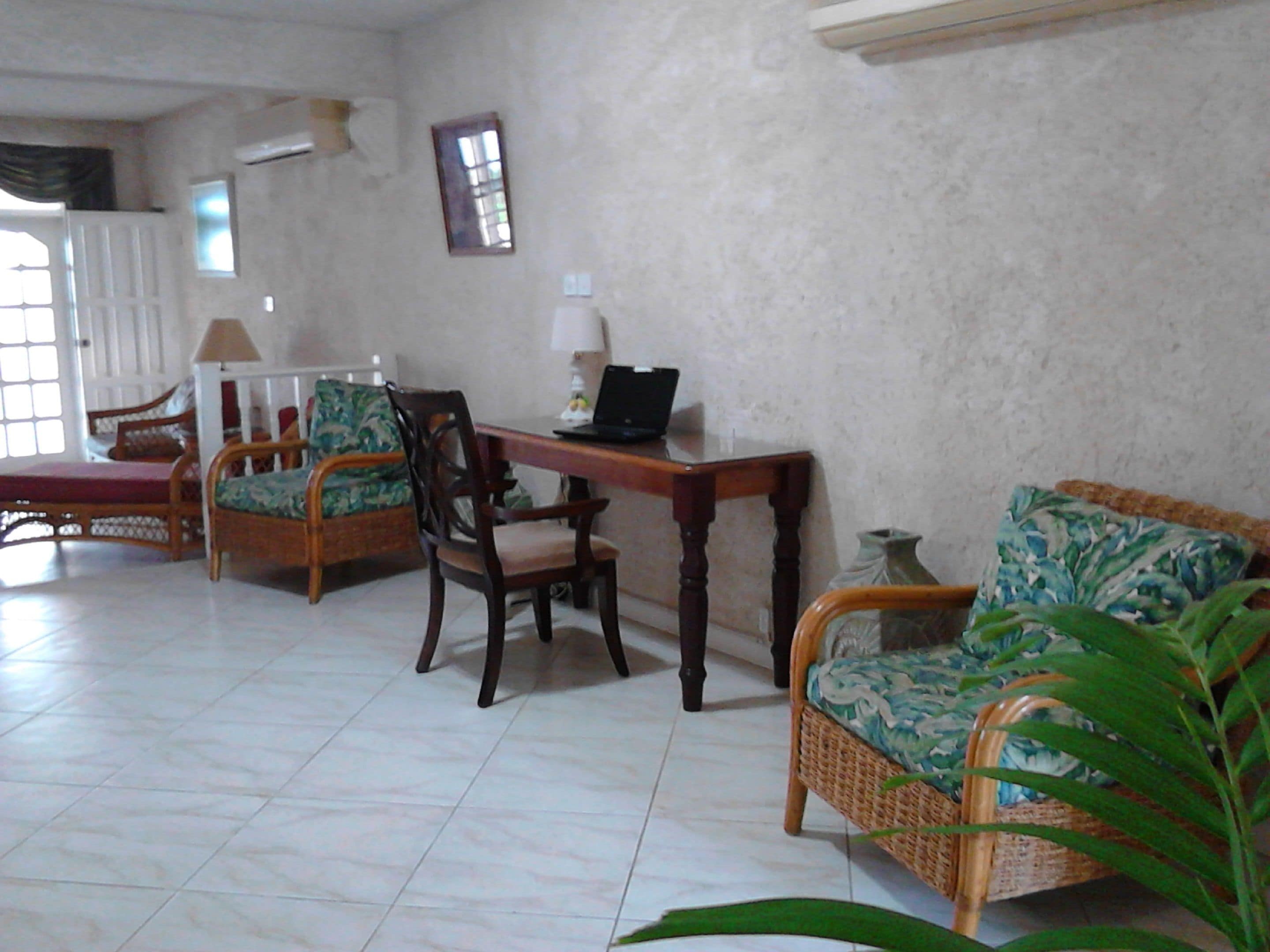 A spacious living area with a tiled floor, a wooden desk with a laptop, a wicker sofa, and two wicker chairs with cushions.