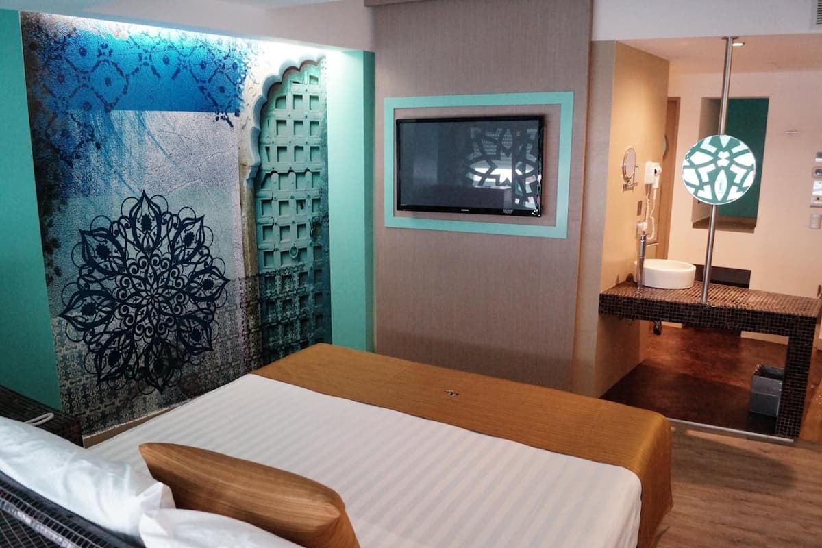 Habitación de hotel con cama, televisor montado en la pared, baño con espejo y lavamanos, y una pared decorativa con motivo geométrico.