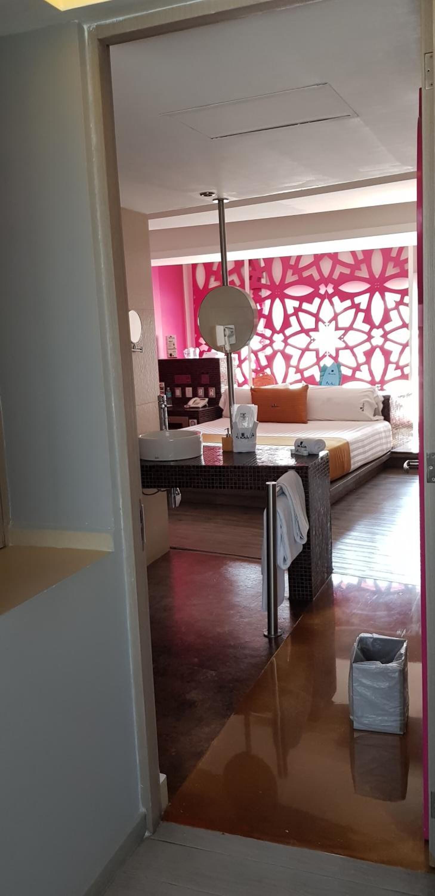 A room with a bed, a bedside table, a mirror, and a wall with a pink and white pattern.