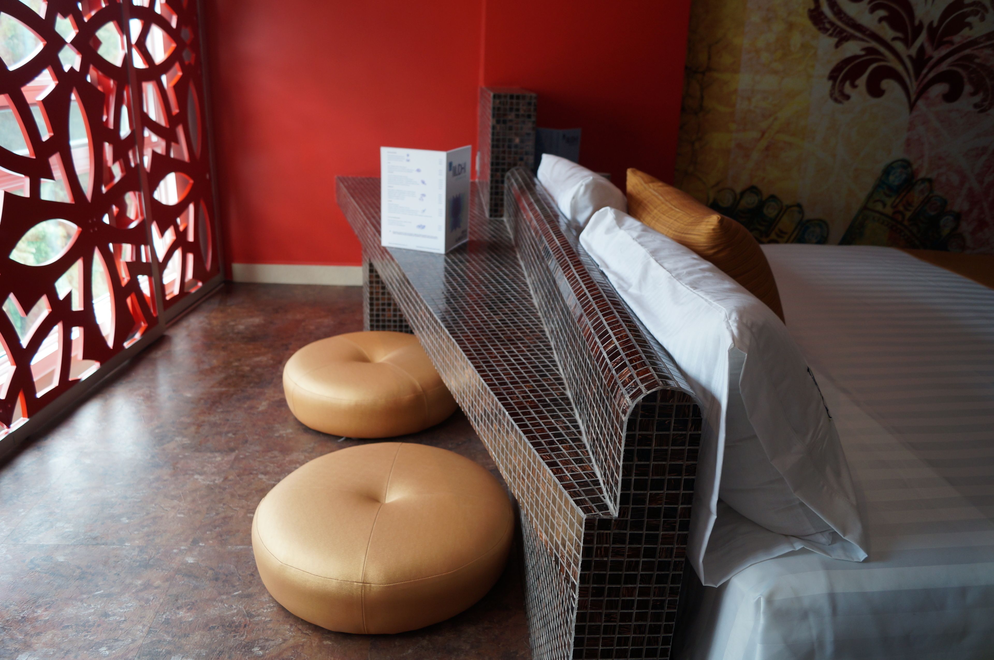 A hotel room with a bed, two round ottomans, a decorative wall, and a red partition.