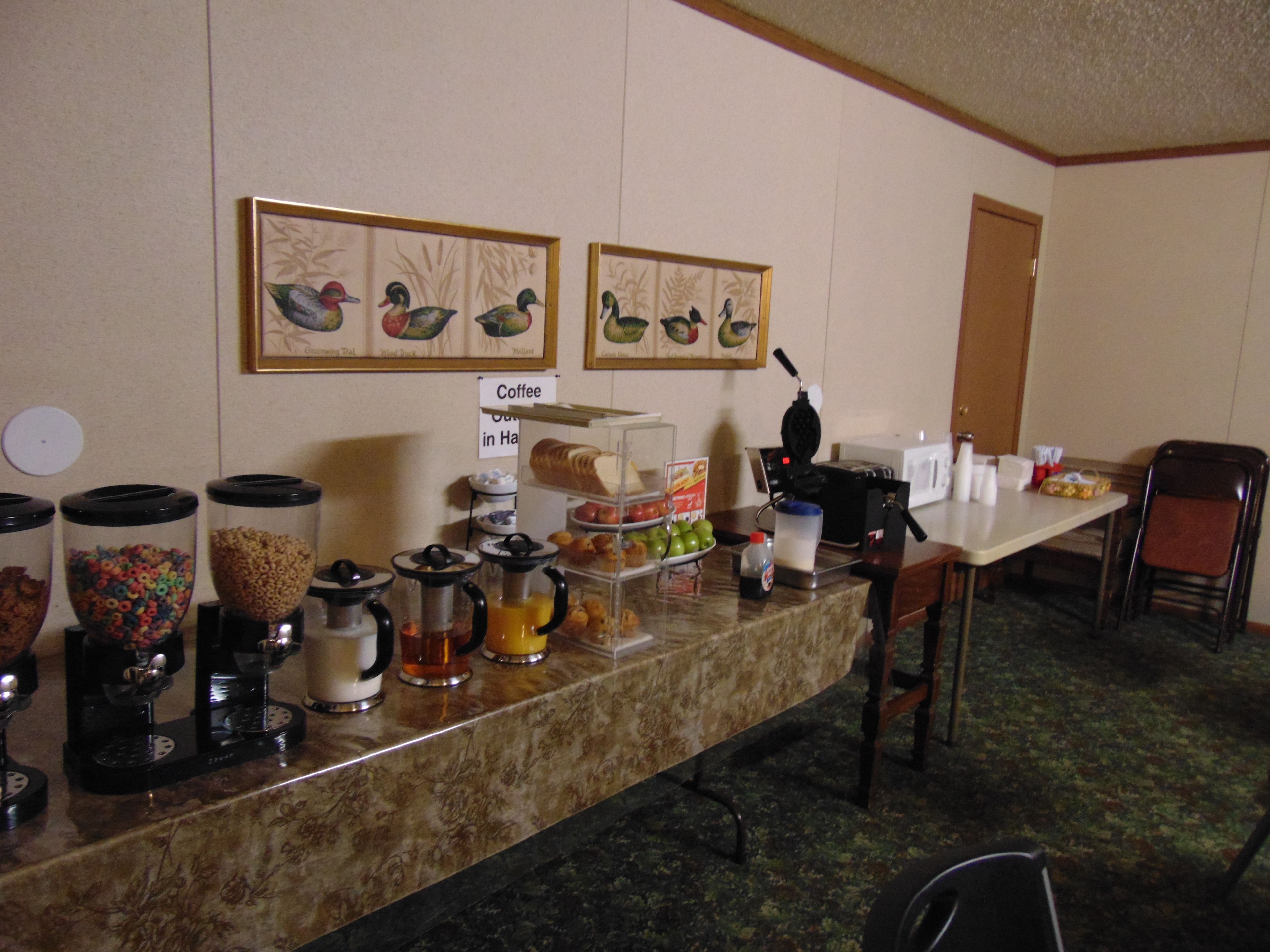 A breakfast bar with various food items and a coffee station. There are framed pictures on the wall and a sign indicating the coffee area.