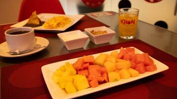 Una mesa de comedor con un plato de fruta en cubos, una taza de café y un vaso de jugo de naranja.