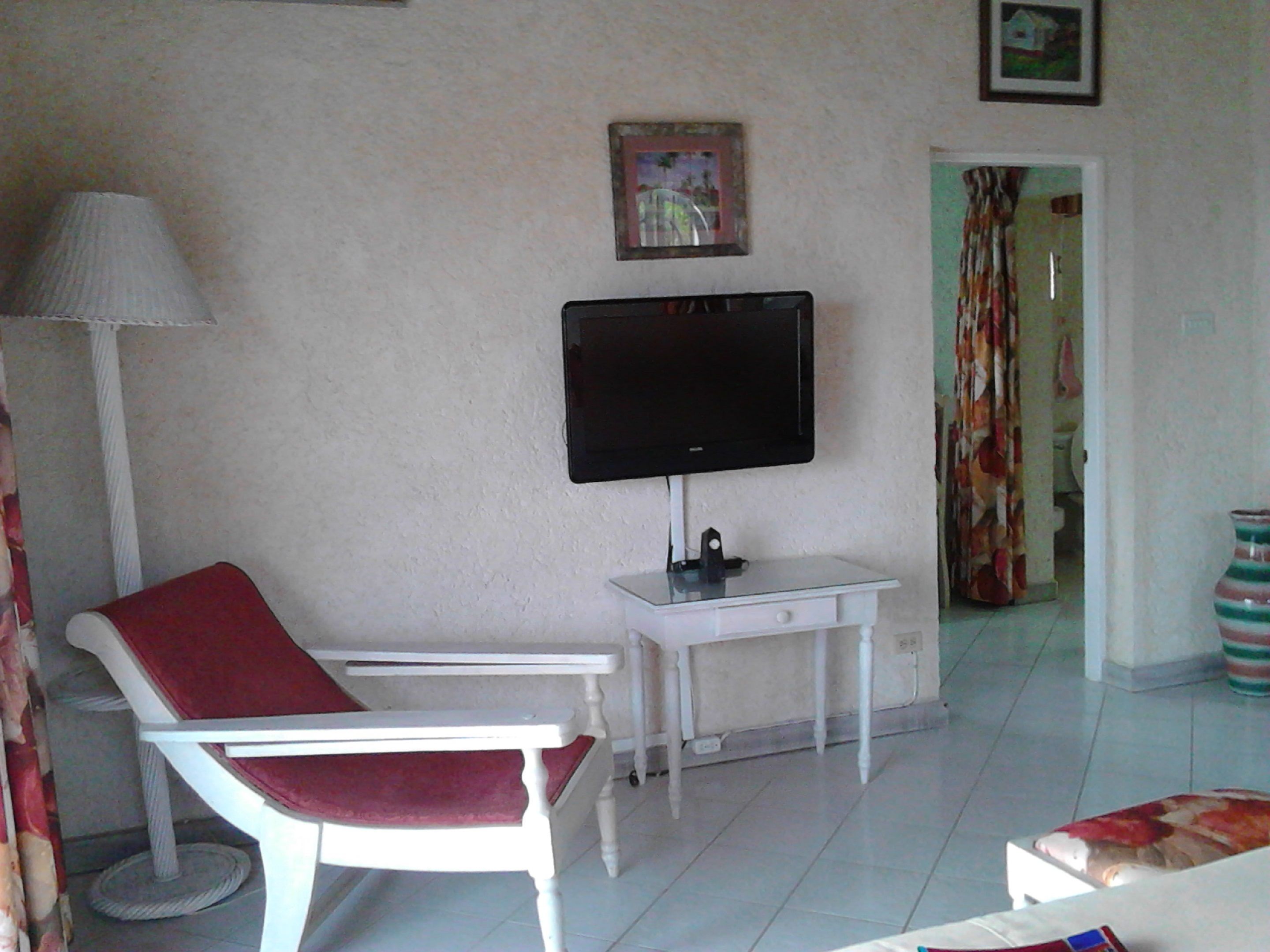 A living room with a white armchair, a television on a stand, a small table, and a door leading to another room.