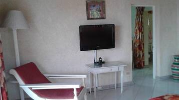 A living room with a white armchair, a television on a stand, a small table, and a door leading to another room.