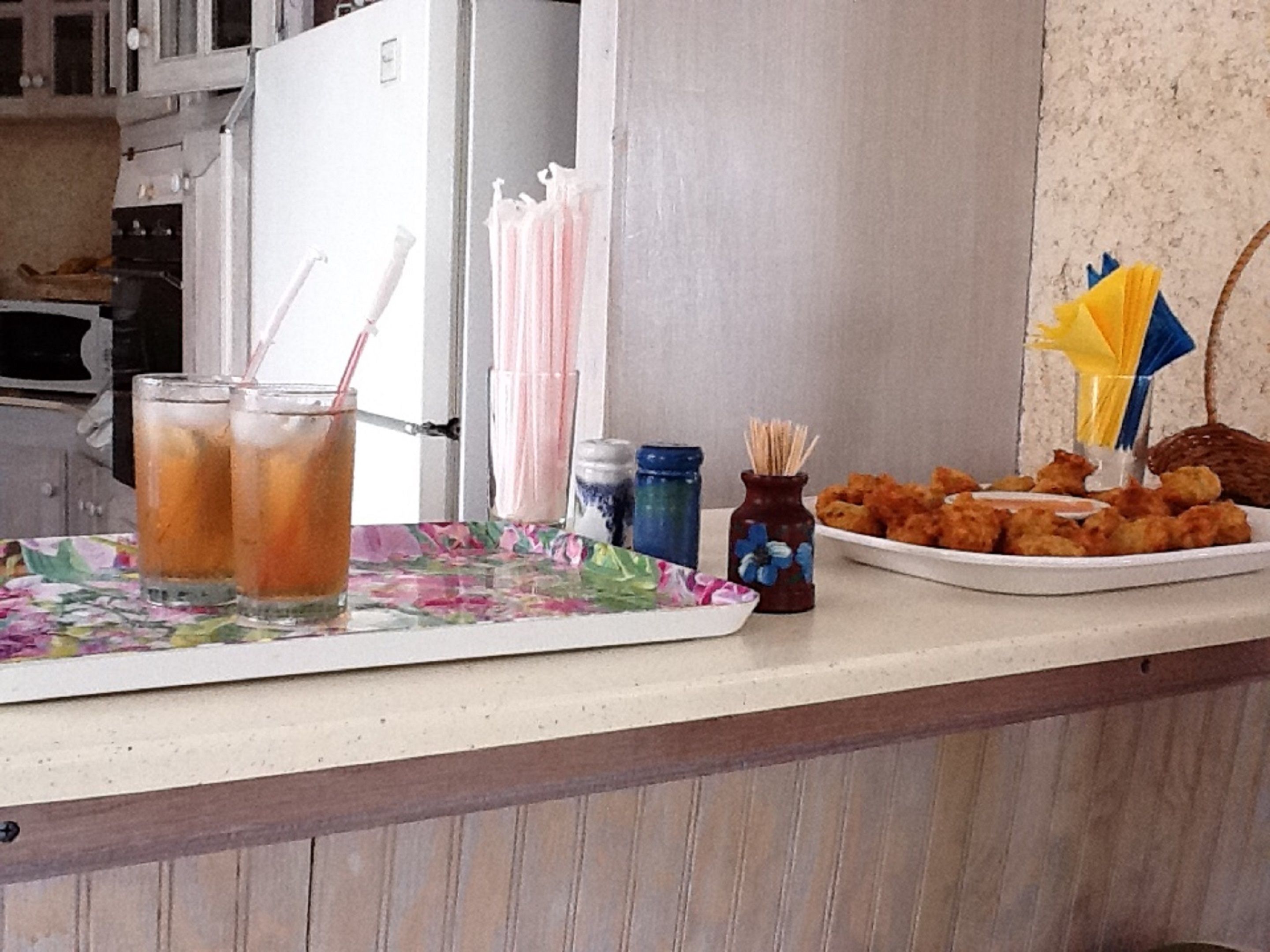 A kitchen counter with a floral tablecloth, two iced drinks, a plate of fried chicken, and a salt shaker.