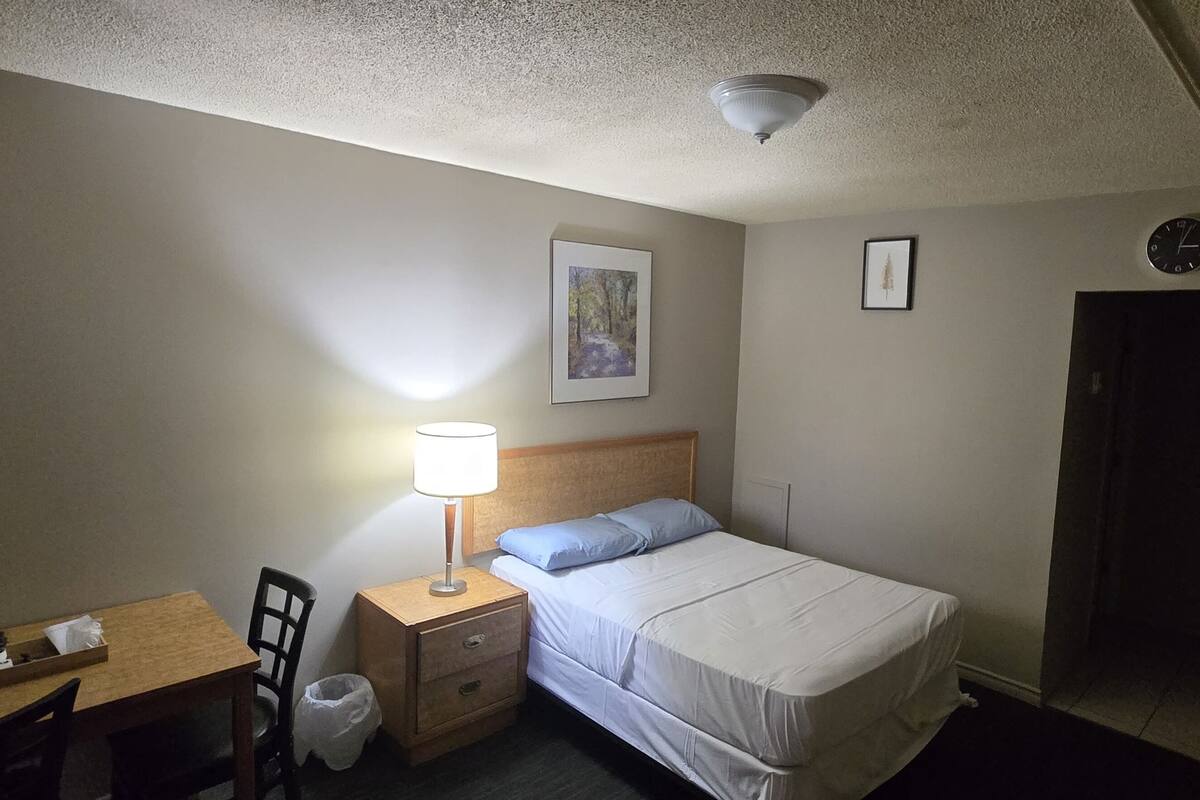 Quarto de hotel com cama, escrivaninha, cadeira, mesa de cabeceira com abajur, lixeira e porta.