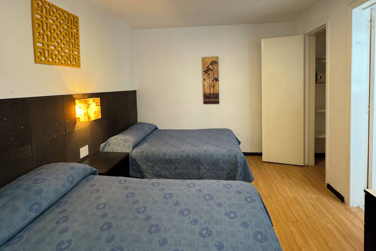 A hotel room with two beds, a wooden floor, a wall-mounted light fixture, and a door leading to another room.