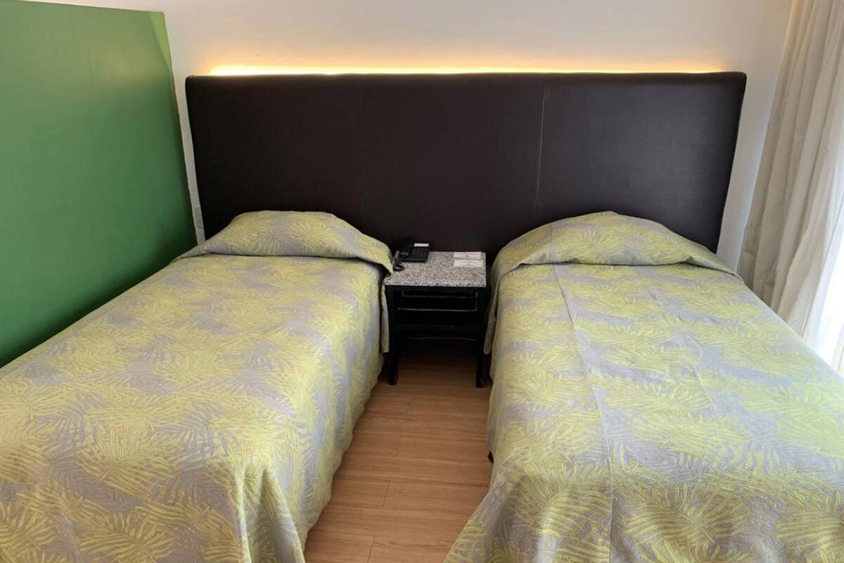 A hotel room with two single beds, a nightstand with a lamp, and a green accent wall.