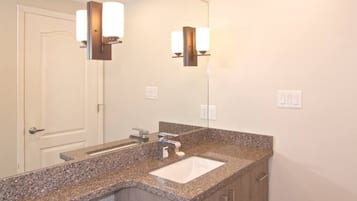 Une salle de bain dotée d’un comptoir en granit, d’un lavabo blanc, d’un miroir et de deux appliques murales.