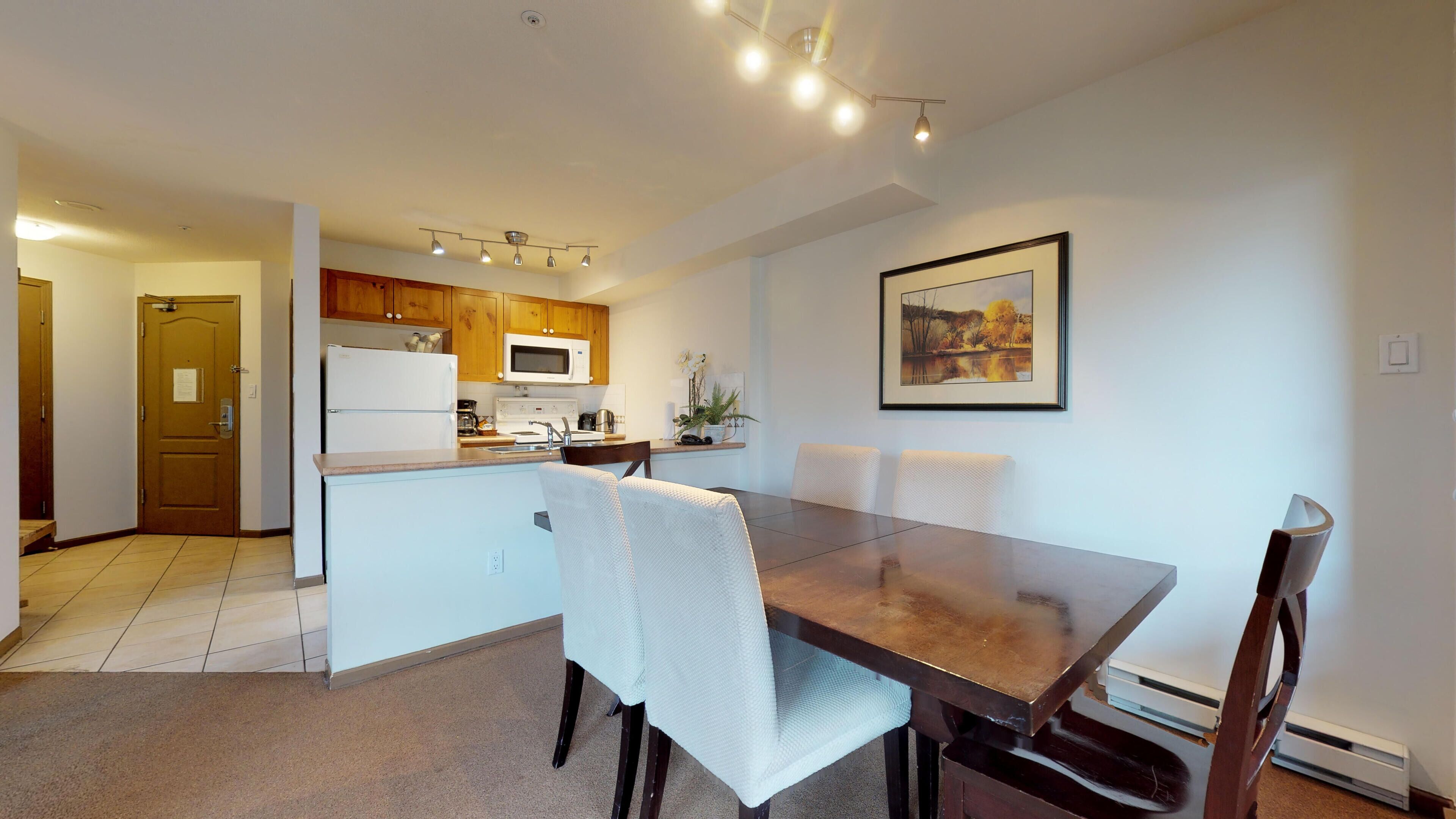 Una cocina moderna con armarios de madera, un refrigerador blanco y un microondas. Hay una mesa de comedor con sillas y un cuadro en la pared.