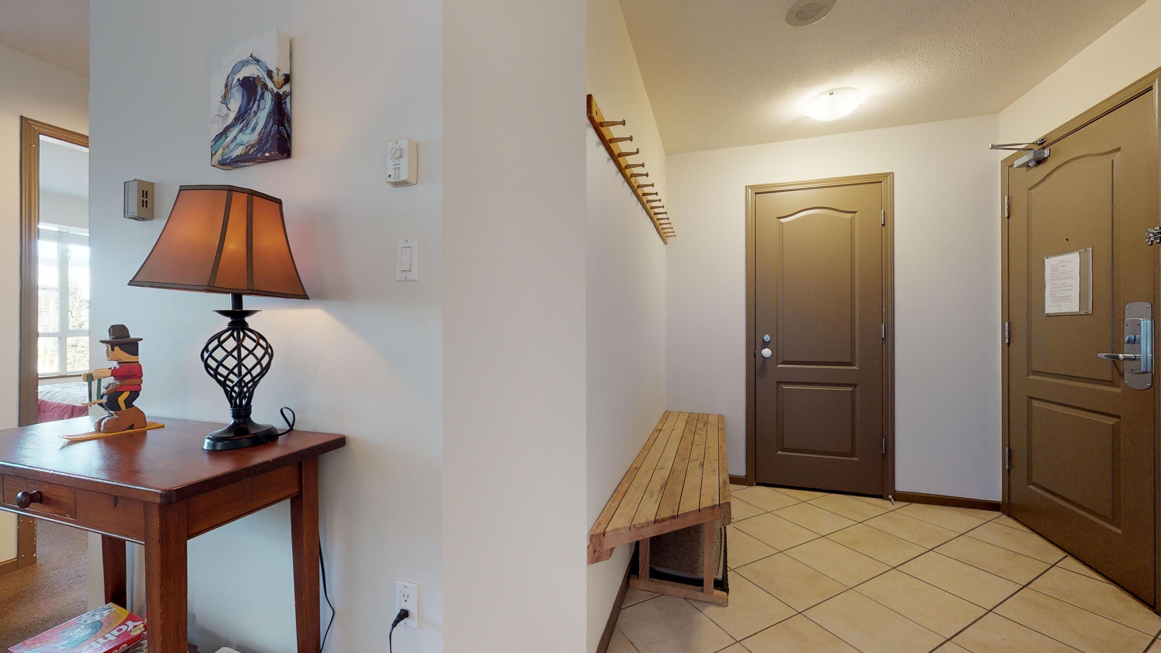 A hallway with a wooden table, a lamp, a small shelf with figurines, a door, and a wall-mounted shelf.