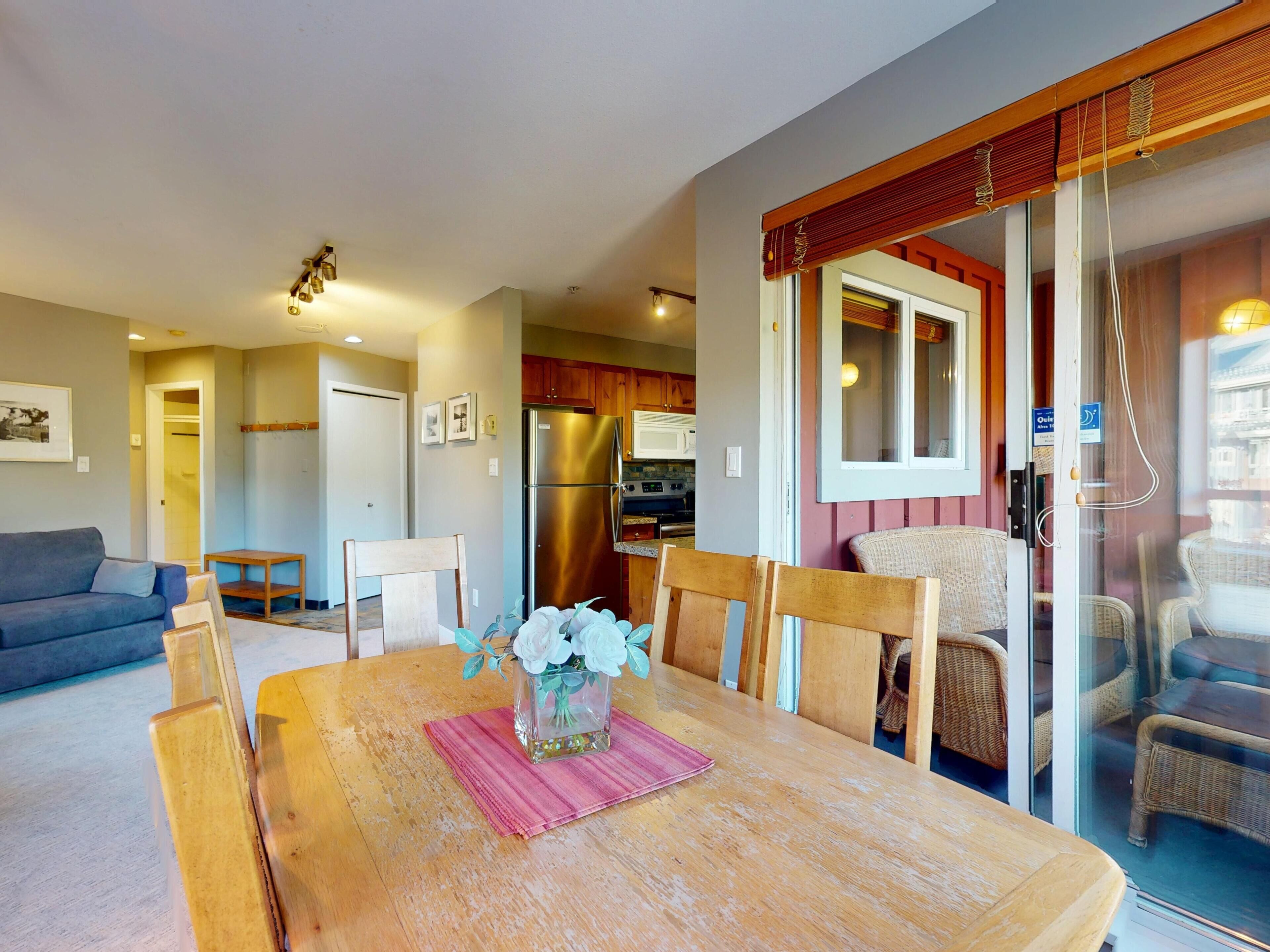 Comedor con mesa de madera, sillas y un florero. Se observa una cocina al fondo, con refrigerador y microondas.