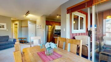 Comedor con mesa de madera, sillas y un florero. Se observa una cocina al fondo, con refrigerador y microondas.