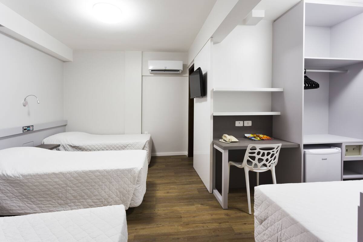 A compact hotel room with two single beds, a desk with a chair, a small refrigerator, and a TV mounted on the wall.