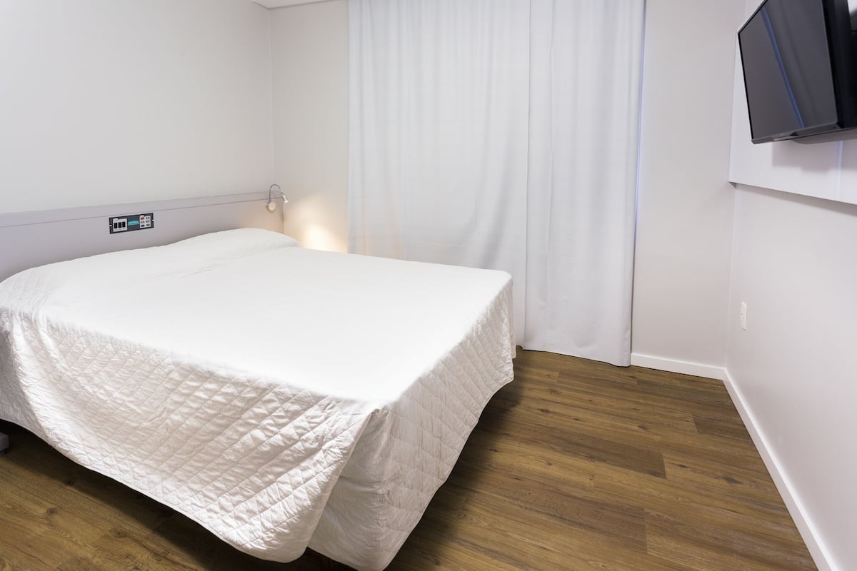 A hotel room with a bed, wooden flooring, a TV mounted on the wall, and a curtain.