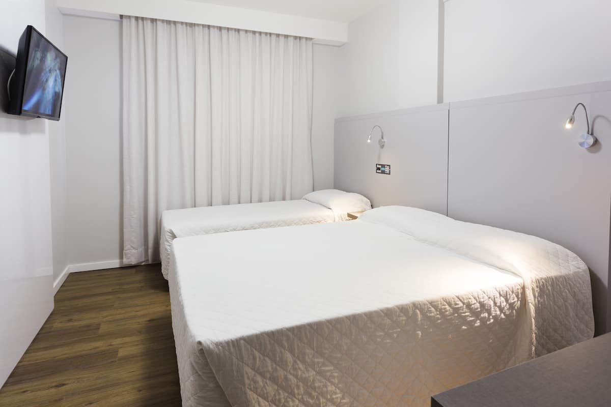A hotel room with two beds, a flat-screen TV mounted on the wall, a bedside table, and a window with curtains.