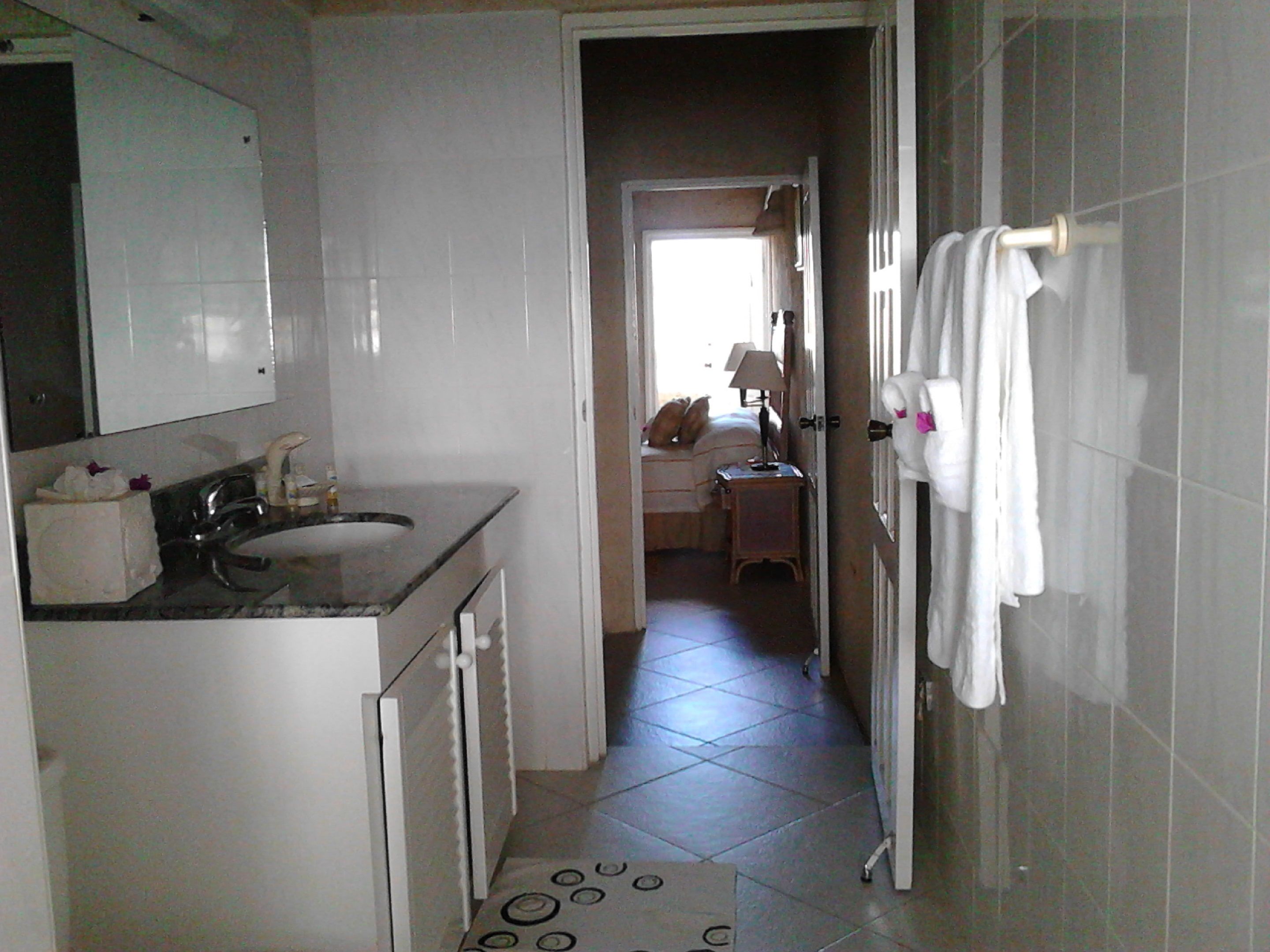 A narrow hallway leading to a bedroom with a bed, a nightstand, and a lamp. A bathroom with a sink and mirror is visible to the left.