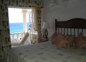 A hotel room with a bed, pillows, a nightstand, a lamp, and a view of the sea through an open door. - Seabliss Villa (Fryers Well)